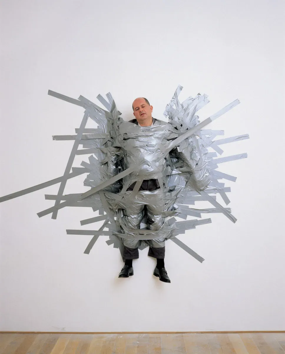 Il gallerista milanese Massimo de Carlo, fissato al muro del suo spazio espositivo con del nastro adesivo da Maurizio Cattelan, per la performance-opera A perfect day (1999).